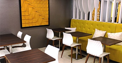 Interior moderno da Pâtisserie Dacquoise com bancos amarelos, cadeiras geométricas brancas, mesas de madeira escura, ripas verticais brancas, plantas verdes exuberantes e uma grande obra de arte abstrata amarela. Bem vindo a um ambiente convidativo da Pastelaria Francesa.