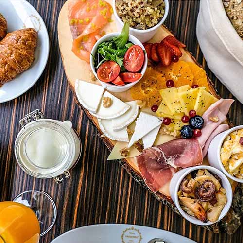 Brunch em Campo de Ourique. Uma travessa de madeira com queijos variados, carnes frias, tomates frescos, salada, fruta fatiada e molhos rodeia os croissants, o sumo de laranja e o leite numa mesa às riscas - perfeito para um brunch Dacquoise descontraído em Lisboa