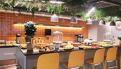 Um balcão de buffet bem iluminado com cadeiras amarelas exibe vários bolos, sanduíches e sobremesas sob cúpulas de vidro - perfeito para catering finger food. O pano de fundo apresenta azulejos cor de laranja, luzes suspensas e uma decoração verdejante por cima.