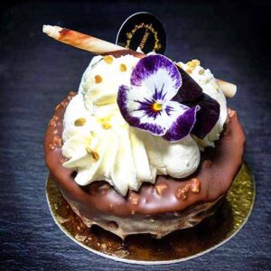 Galeria. Os bolos da Pâtisserie Dacquoise. Uma sobremesa decadente dos bolos da Pâtisserie Dacquoise, coberta com chantilly, um pau de chocolate, granulado dourado e uma flor comestível roxa e branca numa base dourada, destaca-se como arte numa galeria contra um fundo escuro.