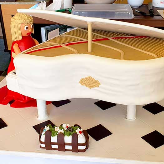 Um bolo com a forma de um piano de cauda com uma mulher de vestido vermelho nas teclas, mais uma pequena sobremesa à frente, é apresentado numa superfície branca com quadrados pretos - uma elegante inspiração de decoração para bolos e coberturas para bolos criativos.