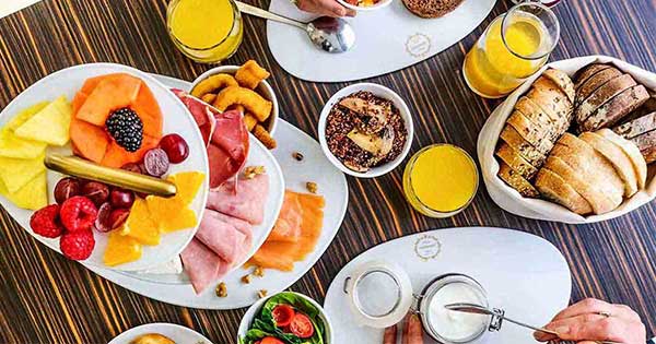 Uma exposição de tabuleiros com fatias de fruta fresca variada, incluindo kiwi, ananás, morangos e melão - perfeitos para um brunch Dacquoise vibrante em Campo de Ourique - juntamente com carnes enroladas e pequenos aperitivos em pratos brancos.