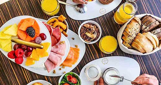 Uma exposição de tabuleiros com fatias de fruta fresca variada, incluindo kiwi, ananás, morangos e melão - perfeitos para um brunch Dacquoise vibrante em Campo de Ourique - juntamente com carnes enroladas e pequenos aperitivos em pratos brancos.