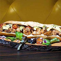 Uma sanduíche comprida com pedaços de frango grelhado, fatias de abacate, tomate cherry e folhas verdes, perfeita para catering para festas particulares, servida numa tábua de madeira rústica com salada e cebolas em cubos à parte.