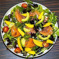 Uma salada colorida com verduras mistas, tomate cereja, salmão fumado, abacate e manga num prato branco sobre uma superfície de madeira escura.