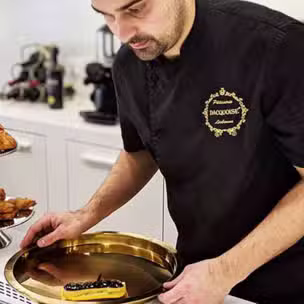 O Chefe Ivo Camões, de uniforme preto, coloca uma sobremesa com chocolate e frutos secos num tabuleiro dourado, numa cozinha moderna.