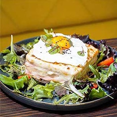 Um prato com um croque madame coberto com um ovo estrelado e microgreens, rodeado por uma salada fresca com verdes mistos e fatias de pimento vermelho - perfeito para catering para aniversários ou festas particulares.