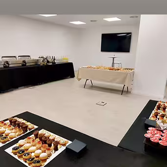 Uma sala moderna e bem iluminada, preparada para um buffet com tabuleiros de pastelaria e sobremesas variadas em cima das mesas e um grande ecrã montado na parede para um ambiente adicional. No seu Catering ao Domicílio