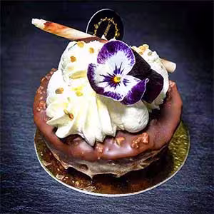 Galeria. Os bolos da Pâtisserie Dacquoise. Uma sobremesa decadente dos bolos da Pâtisserie Dacquoise, coberta com chantilly, um pau de chocolate, granulado dourado e uma flor comestível roxa e branca numa base dourada, destaca-se como arte numa galeria contra um fundo escuro.