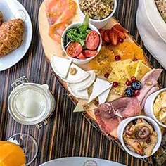 Uma bandeja de madeira com queijos variados, carnes curadas, bagas frescas e legumes - perfeita para catering para aniversários ou festas particulares - rodeada por pastas de barrar, um croissant, sumo de laranja e leite numa mesa às riscas.