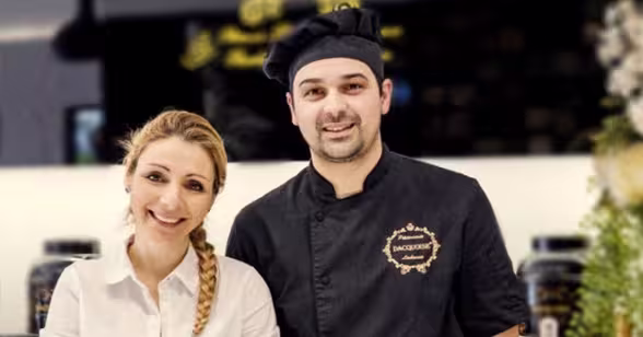 Uma mulher sorridente e o Chefe Ivo Camões estão atrás de um balcão de pastelaria. Um letreiro preto e dourado de uma pastelaria é visível ao fundo, dando um vislumbre de quem somos.