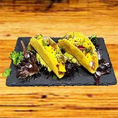 Dois tacos de casca dura recheados com alface, guacamole, manga cortada e cebola roxa em cubos, servidos num prato de ardósia preta com mistura de verduras, tudo muito bem apresentado numa superfície de madeira.