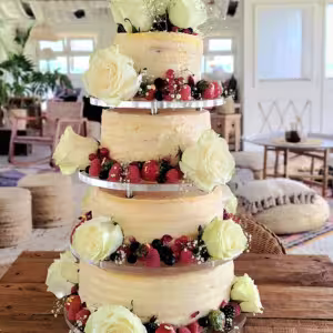 Naked cake de três andares, adornado com grandes rosas brancas, bagas e pequenas flores brancas - exposto numa mesa de madeira num ambiente interior acolhedor e rústico.