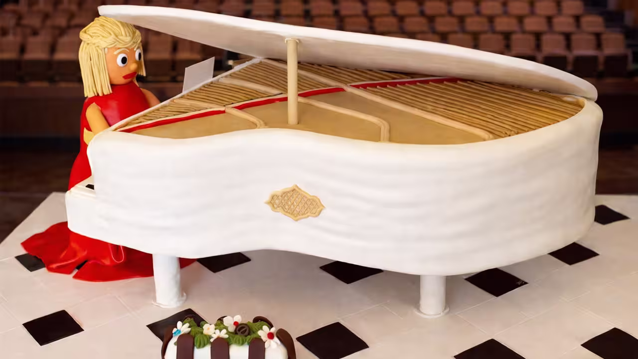 Um bolo com a forma de um piano de cauda com uma mulher de vestido vermelho nas teclas, mais uma pequena sobremesa à frente, é apresentado numa superfície branca com quadrados pretos - uma elegante inspiração de decoração para bolos e coberturas para bolos criativos.