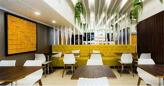 Interior moderno da Pâtisserie Dacquoise com bancos amarelos, cadeiras geométricas brancas, mesas de madeira escura, ripas verticais brancas, plantas verdes exuberantes e uma grande obra de arte abstrata amarela. Bem vindo a um ambiente convidativo da Pastelaria Francesa.