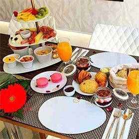 Uma mesa de pequeno almoço posta para um brunch em Campo de Ourique, Lisboa, com pastelaria variada, macarons, fruta fresca, carnes frias, queijo, taças de cereais, sumo de laranja, um centro de mesa de flores vermelhas e duas cadeiras brancas ao fundo.
