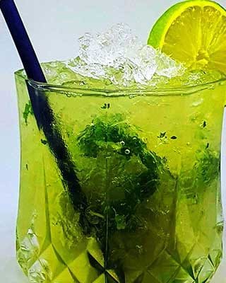 Um copo de cocktail caipirinha com gelo picado, folhas de hortelã, uma rodela de lima e uma palhinha escura, servido num copo facetado contra um fundo liso.