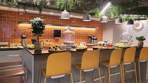 Um balcão de buffet bem iluminado com cadeiras amarelas exibe vários bolos, sanduíches e sobremesas sob cúpulas de vidro - perfeito para catering finger food. O pano de fundo apresenta azulejos cor de laranja, luzes suspensas e uma decoração verdejante por cima.