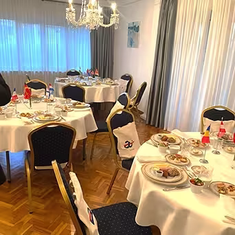 Uma sala de jantar com mesas redondas preparadas para uma refeição, cada uma com pratos de comida, copos e pequenas bandeiras internacionais - perfeito para coffee break catering Lisboa. Cadeiras azuis almofadadas rodeiam as mesas por baixo de um lustre cintilante.