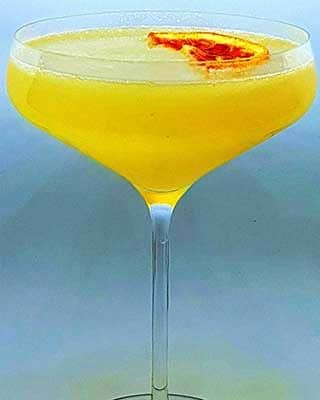 Um cocktail amarelo num copo coupé, decorado com uma pequena rodela de laranja seca, perfeito para festas particulares e eventos sofisticados, sobre um fundo claro e simples.