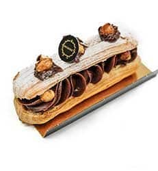 Um éclair recheado com um rico creme de chocolate, coberto com avelãs, decorações de chocolate, açúcar em pó e uma etiqueta redonda das nossas Coleções Sazonais, apresentado numa tábua retangular dourada sobre um fundo branco.