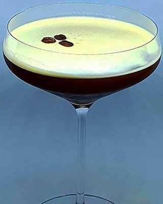 Um copo transparente cheio de um martini expresso escuro, coberto com espuma cremosa e três grãos de café, perfeito para catering para festas particulares, sobre um fundo claro e simples.