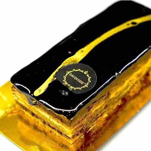 Um Bolo Opéra retangular com uma cobertura de chocolate preto brilhante, decorado com uma faixa diagonal dourada e um rótulo redondo, colocado elegantemente numa bandeja dourada.