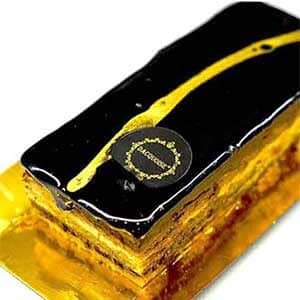 Um Bolo Opéra retangular com uma cobertura de chocolate preto brilhante, decorado com uma faixa diagonal dourada e um rótulo redondo, colocado elegantemente numa bandeja dourada.