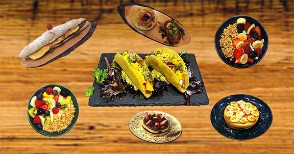Uma mesa de madeira exibe vários pratos: tacos numa travessa de ardósia no centro, rodeados por taças de fruta e granola, uma tarte de sobremesa, Saladas dacquoise, mousse de chocolate, um crepe e um prato com uma massa com cobertura de fruta.