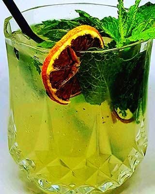 Um copo de mojito com uma palhinha preta, decorado com folhas de hortelã fresca e duas rodelas de lima seca, apresentado sobre um fundo claro.