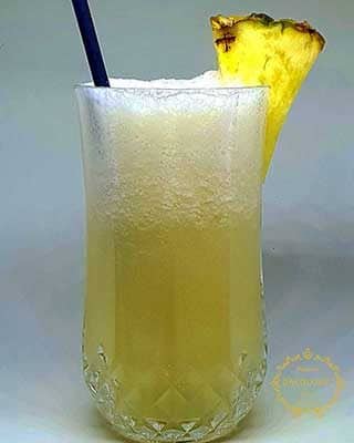 Uma bebida amarela espumosa num copo transparente e texturizado com uma palhinha azul e uma rodela de ananás na borda - perfeita para catering para aniversários, com um fundo claro.