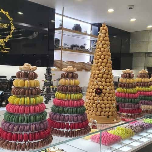 Torres de macarons coloridos e um croquembouche alto estão expostos numa pastelaria, rodeados por prateleiras de frascos e pratos, perfeitos para quem procura bolos de aniversário de crianças ou deliciosos bolos de frutas.