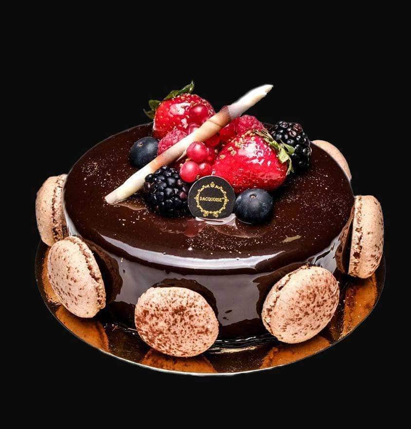 Um bolo de chocolate brilhante coberto com bagas variadas, dois palitos de chocolate e uma pequena etiqueta redonda de contacto. Decorado com macarons nas laterais e com um delicado dacquoise, assenta numa base dourada sobre um fundo preto.