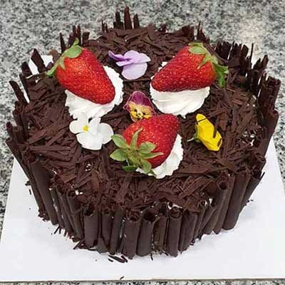 Bolo redondo de chocolate com recheio rico para bolos, decorado com raspas de chocolate, três morangos sobre chantilly, flores comestíveis e caracóis de chocolate nas laterais, colocado sobre uma superfície branca.