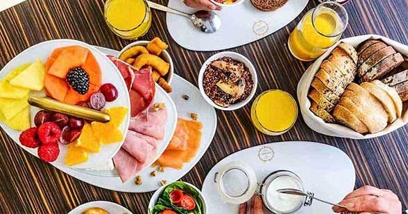 Uma exposição de tabuleiros com fatias de fruta fresca variada, incluindo kiwi, ananás, morangos e melão - perfeitos para um brunch Dacquoise vibrante em Campo de Ourique - juntamente com carnes enroladas e pequenos aperitivos em pratos brancos.