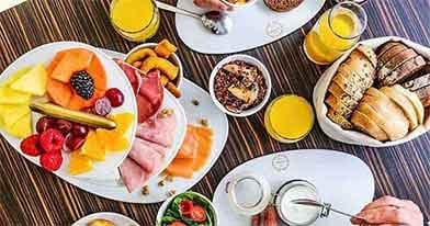 Uma exposição de tabuleiros com fatias de fruta fresca variada, incluindo kiwi, ananás, morangos e melão - perfeitos para um brunch Dacquoise vibrante em Campo de Ourique - juntamente com carnes enroladas e pequenos aperitivos em pratos brancos.
