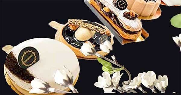 Uma seleção de pastelaria elegante e moderna das nossas Coleções Sazonais com acabamentos brilhantes e logótipos decorativos de chocolate, dispostos sobre um fundo escuro com flores brancas em primeiro plano.