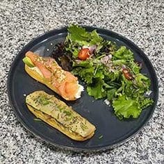 Um prato preto com uma salada verde mista, um éclair com salmão fumado e queijo creme e outro com uma pasta verde salgada, numa bancada salpicada.